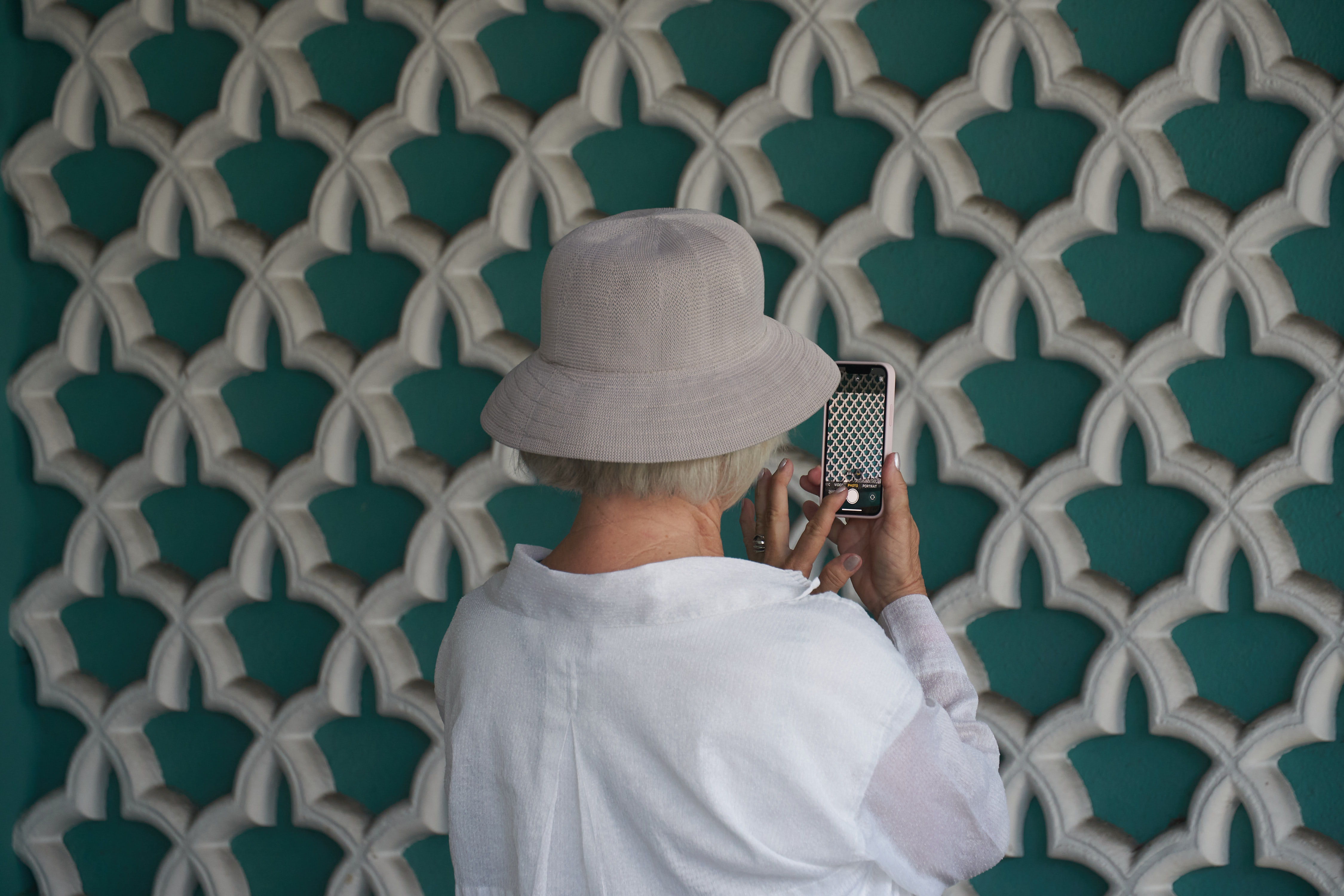 A woman taking a picture of a patterned wall with her cell phone.